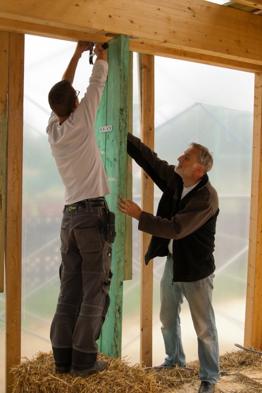 Jens en Papa Ronny zetten de balk op de juiste afstand perfect waterpas recht tussen de balken van het houtskelet. Zo wordt de muur in twee delen gesplitst en kunnen we deze twee delen met stro opvullen.